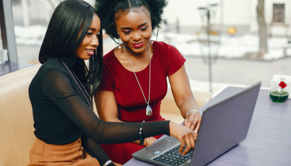 two beautiful and stylish young, dark girls sitting in a restaurant at the table, chatting and using a laptop